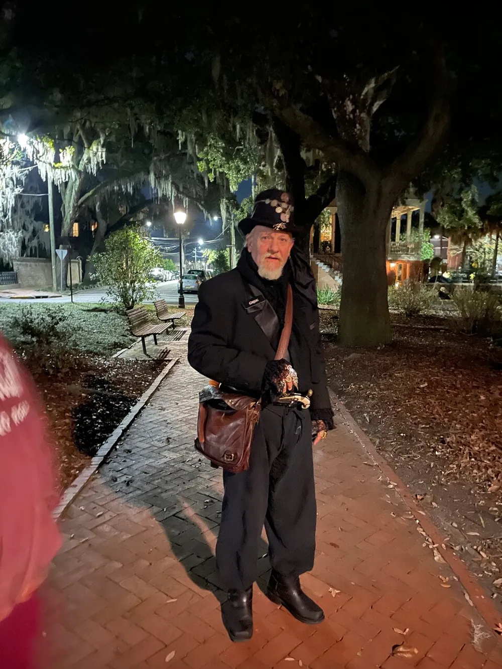 A person dressed in Victorian-era attire is standing on a brick path at night with Spanish moss-draped trees in the background