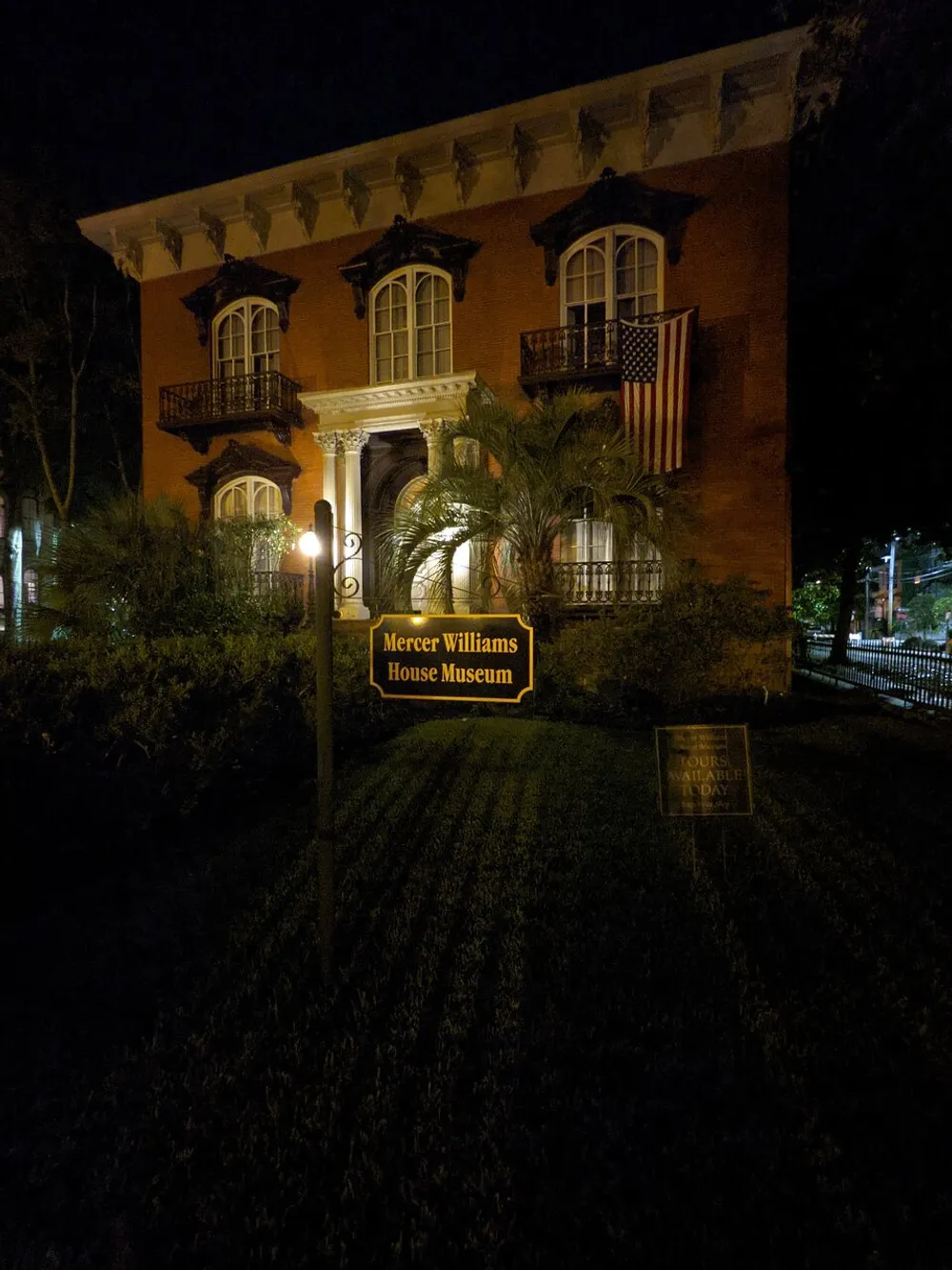 The image shows the exterior of the Mercer Williams House Museum at night illuminated by lamplight and adorned with an American flag with signage in the foreground indicating tours are available