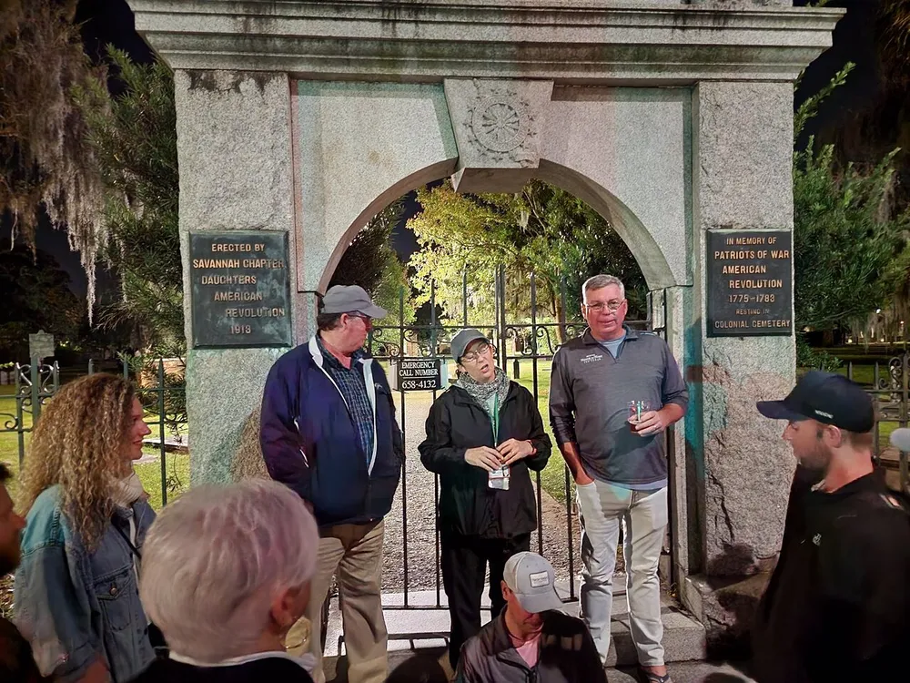 A group of people are gathered at night by an archway entrance to Colonial Cemetery with plaques commemorating the American Revolution on either side