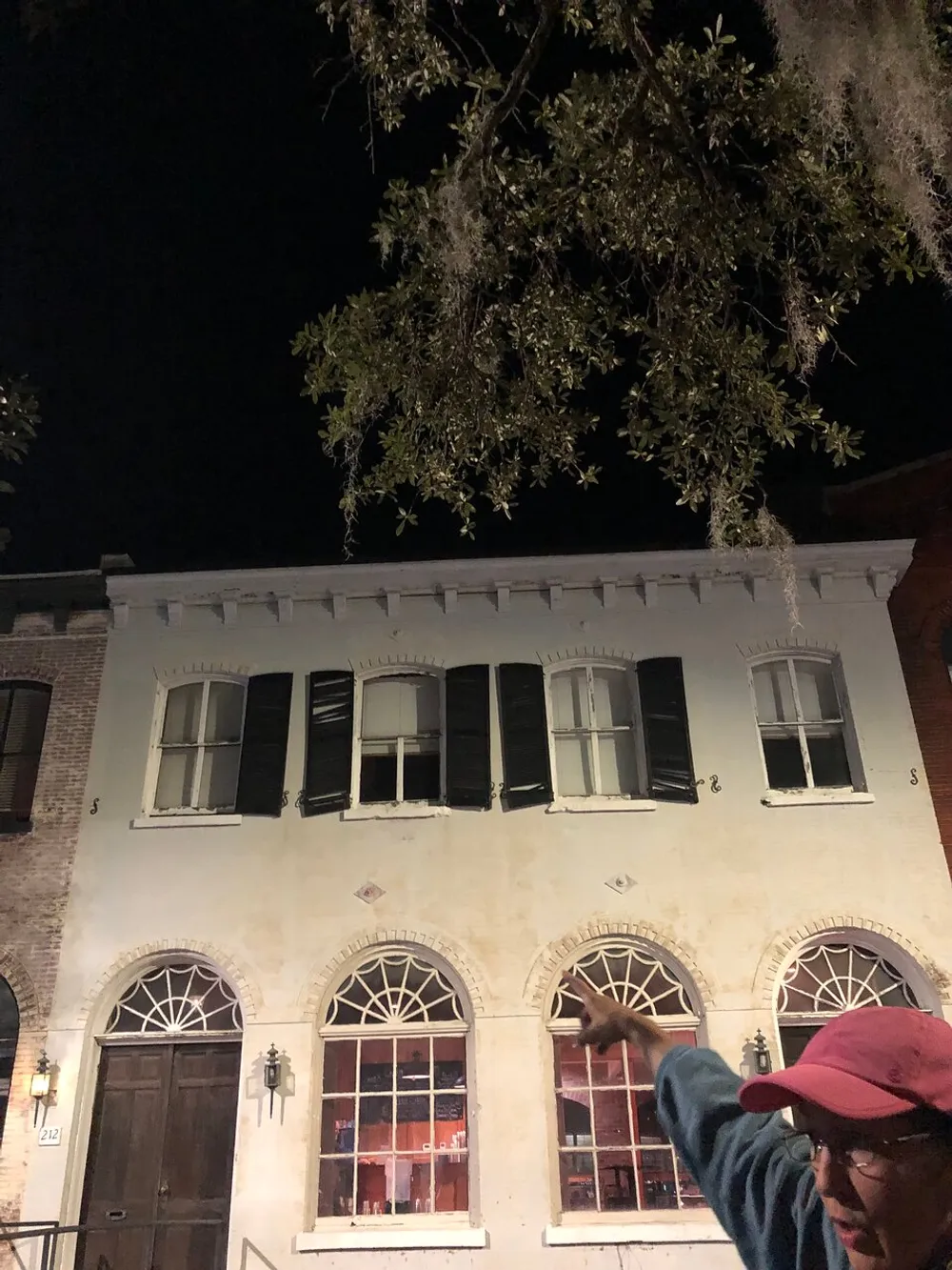 A person in a red hat is gesturing towards a two-story building with black shutters at night under a tree with hanging Spanish moss