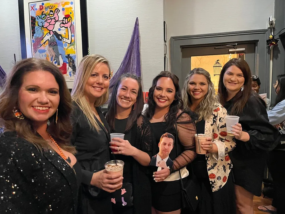 A group of six smiling women are posing together for a photo at an indoor gathering with the woman in the center holding a drink with a cutout of a mans face on it