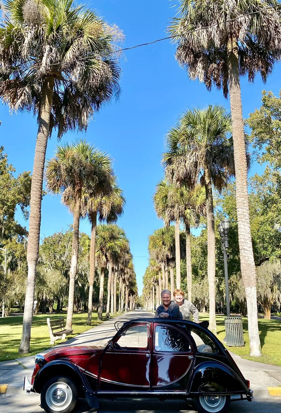 Two people are smiling and popping their heads through the open sunroof of a classic red and black car, parked under a row of palm trees on a sunny day.