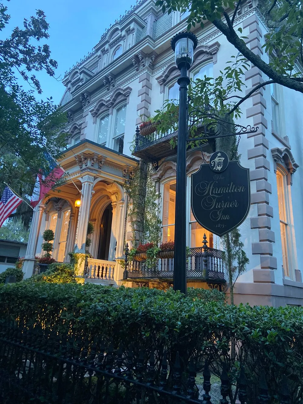 The image shows the exterior of the Hamilton-Turner Inn an elegant historic building adorned with an American flag at twilight with external lighting illuminating the structures detailed architecture