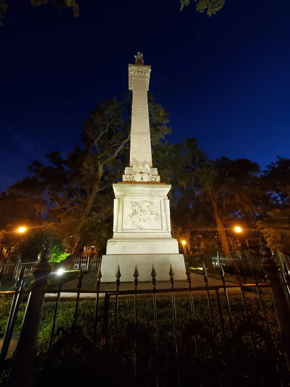 The image depicts a monument illuminated by artificial lighting against a dusky or night sky encircled by a wrought iron fence nestled amongst trees
