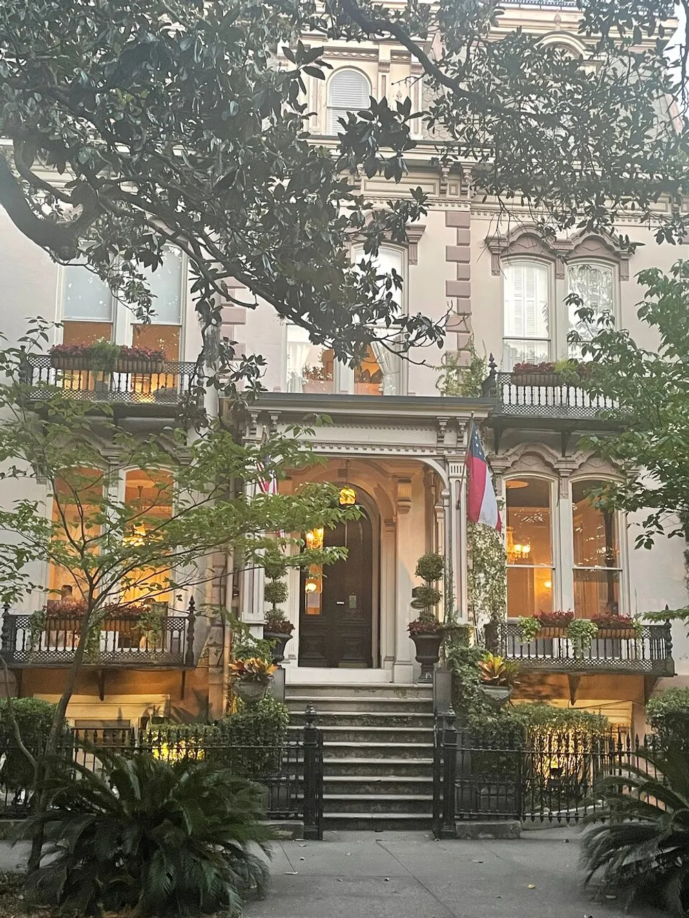 The image shows a picturesque historic building with a warm glow from the lights inside adorned with greenery and flags and framed by surrounding trees and foliage