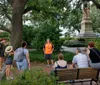 A group of people is listening to a guide wearing an orange shirt in a shaded park with lush greenery and a monument in the background