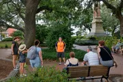 A group of people is listening to a guide wearing an orange shirt in a shaded park with lush greenery and a monument in the background.