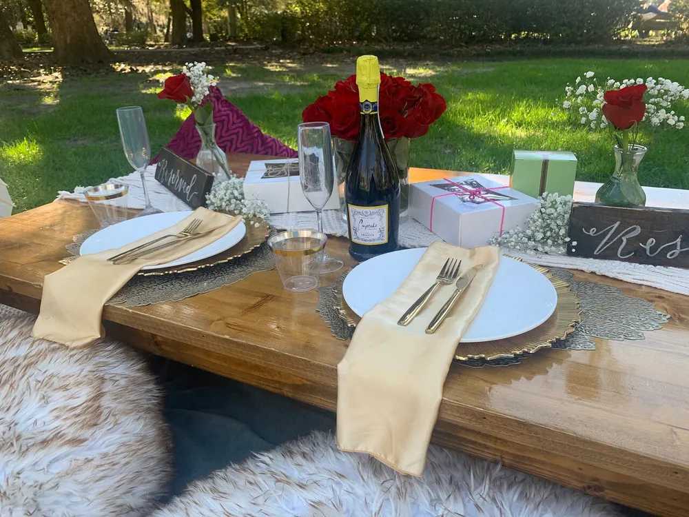 A romantic outdoor picnic setup featuring an elegant table with plates silverware champagne flowers and gifts suggesting a special occasion