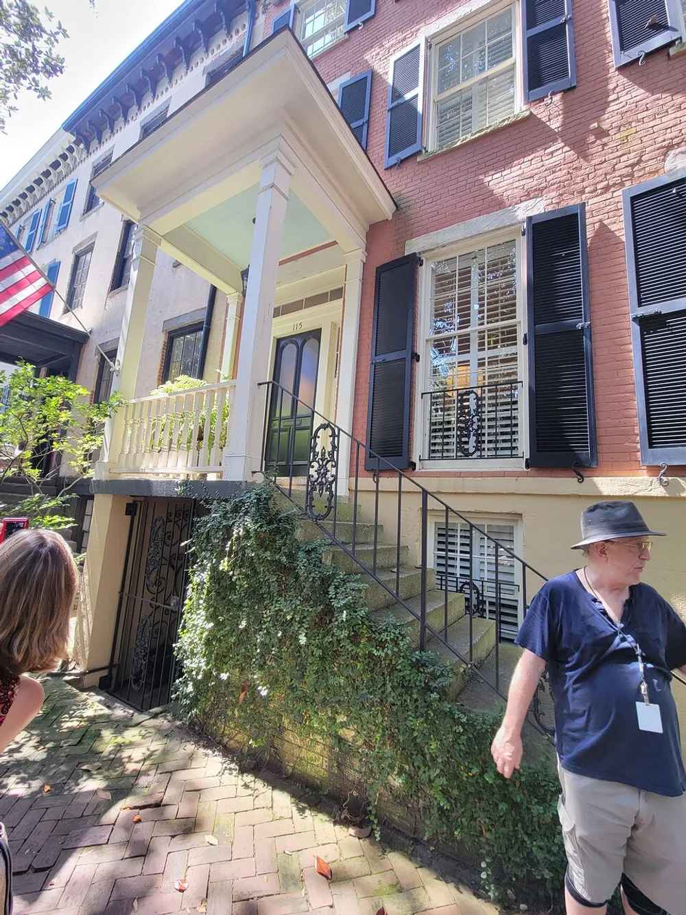 A person is walking by a traditional brick building with black shutters a small front porch and an American flag