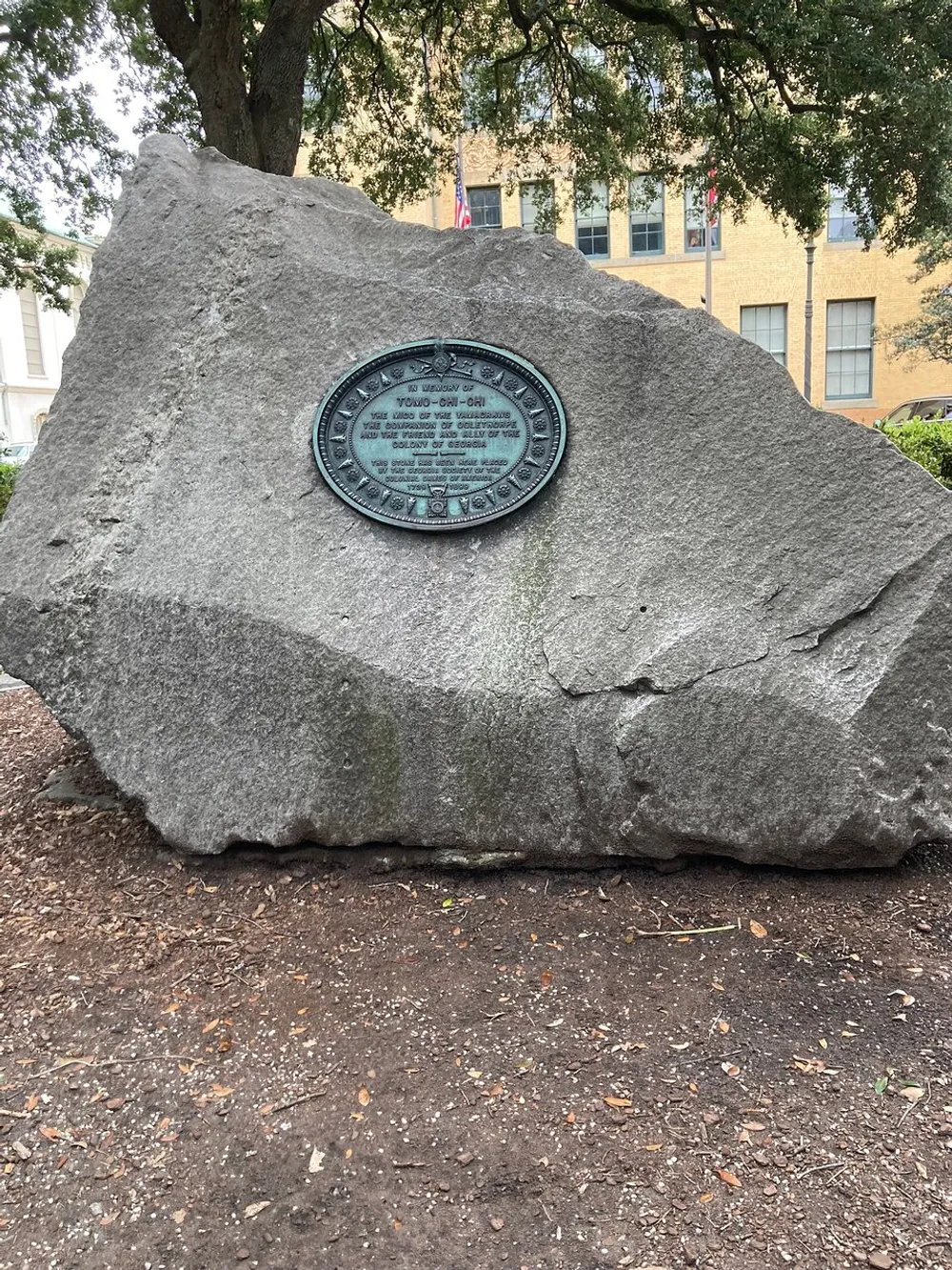 The image shows a large rock with a circular historical marker plaque attached to it located in an outdoor setting with trees and a building in the background