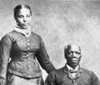 The image is a grainy monochromatic historical photograph depicting two individuals a standing woman in a buttoned-up garment and a seated man in a collared shirt