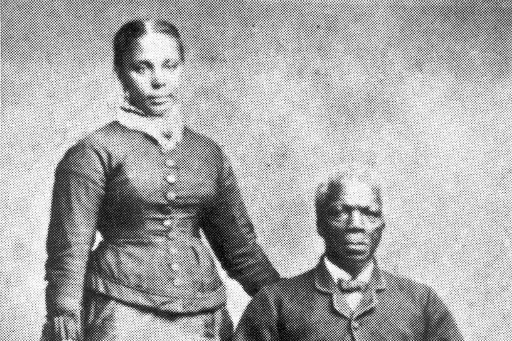 The image is a grainy monochromatic historical photograph depicting two individuals a standing woman in a buttoned-up garment and a seated man in a collared shirt