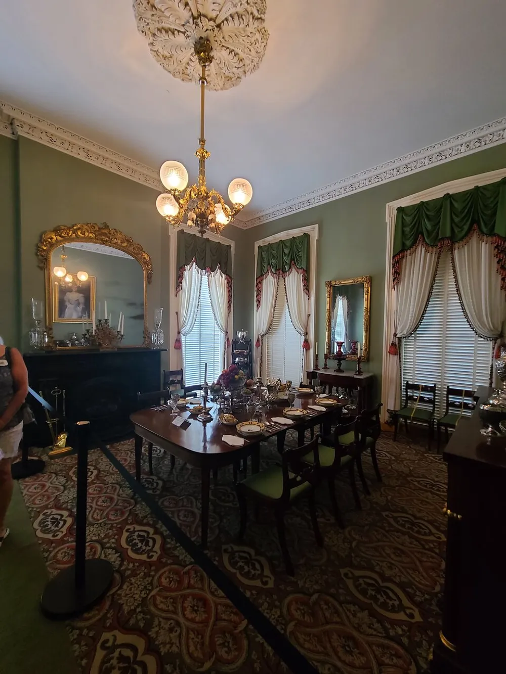 The image showcases an elegant Victorian-style dining room with a chandelier ornate ceiling rose patterned carpet a large mirror restored window draperies and a formally set dining table reflecting historical interior design