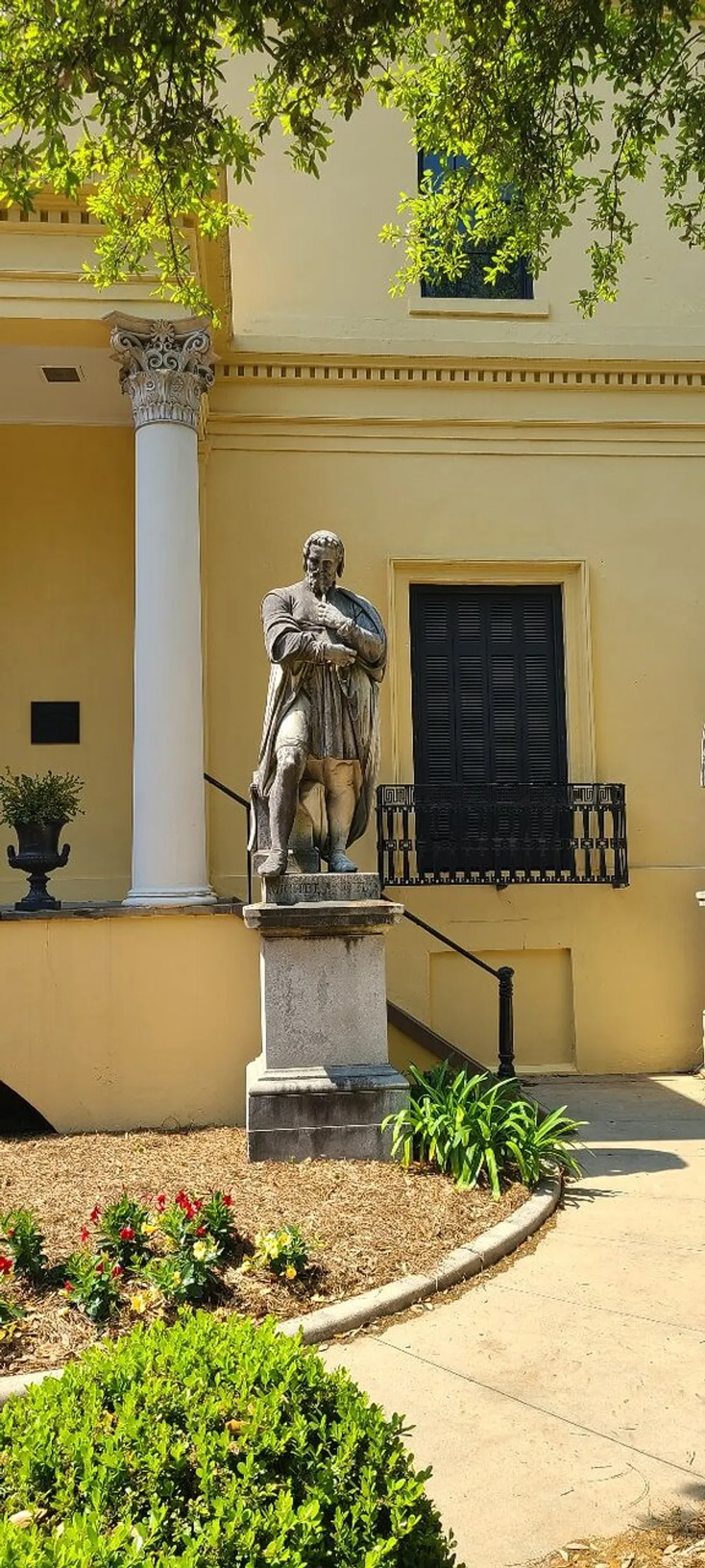 The image shows a stone statue of a bearded figure with a contemplative expression standing in front of a classical building with a column a balcony and greenery