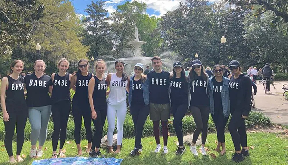 A group of people most wearing matching tops with BABE and one with BRIDE are posing for a photo in a sunny park with a fountain in the background suggesting a bachelorette party
