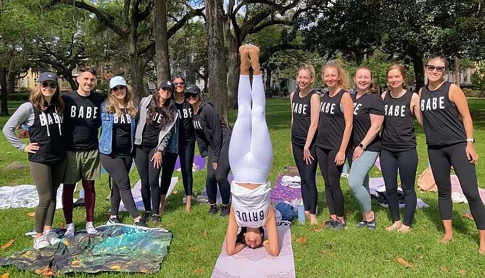 A group of people wearing matching BABE shirts stand in a park with one person doing a headstand in the center