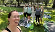 A group of six smiling women are posing for a selfie in a sunny park, probably after a workout or yoga session, with exercise mats on the grass.