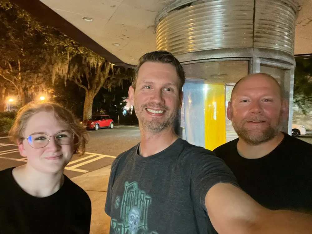 Three people are posing for a smiling selfie at night with trees in the background and a unique cylindrical structure to the right