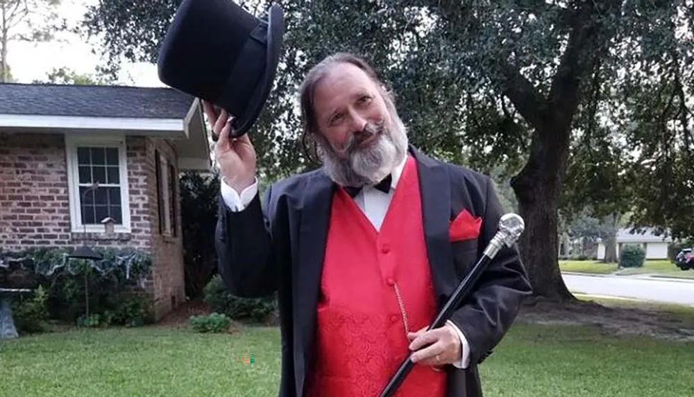 A man with a beard is tipping his top hat while dressed in a formal suit with a red vest and holding a cane