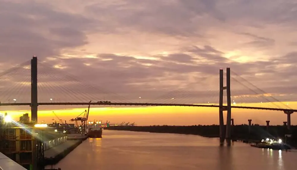 A cable-stayed bridge spans a river with a radiant sunset in the background and industrial structures along the riverbank