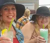 Two women in wide-brimmed hats are smiling with drinks in their hands at an outdoor venue