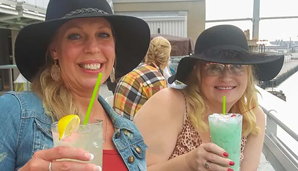 Two women in wide-brimmed hats are smiling with drinks in their hands at an outdoor venue
