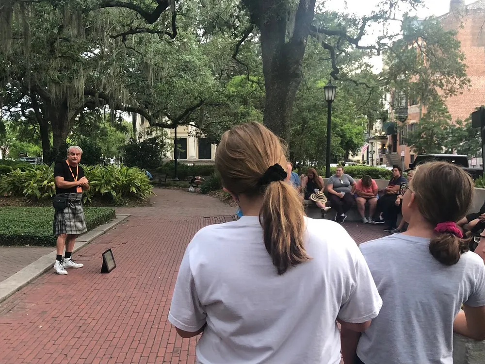 A group of people attentively listens to an individual speaking outdoors in a park-like setting with trees draped in Spanish moss