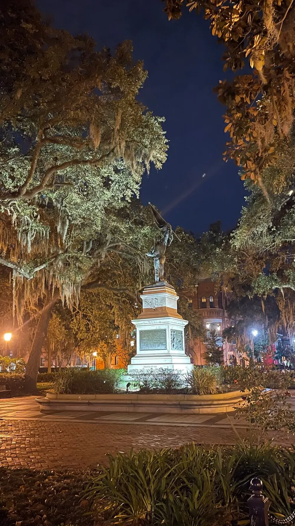 The image depicts a nighttime scene of a statue on a pedestal surrounded by a lush garden with overhanging trees draped in Spanish moss illuminated by nearby streetlamps