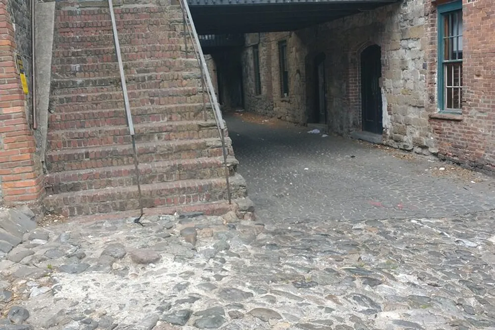 The image shows a cobblestone ground leading to an underpass with old brick walls and a staircase on the left