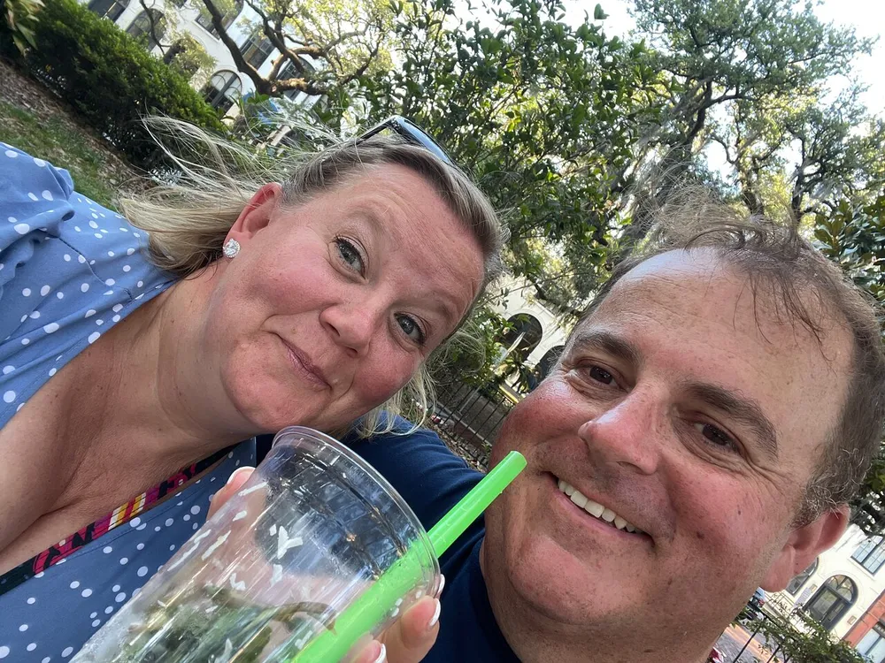 Two people are posing for a close-up selfie outdoors with trees in the background and one of them is holding a drink with a green straw