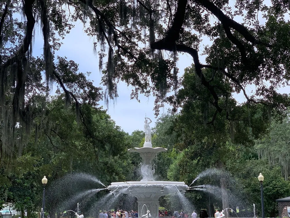 The image captures a picturesque fountain surrounded by lush trees draped with Spanish moss with people casually gathering around it