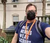 A person is taking a selfie while wearing a mask glasses a T-shirt that reads REAL HEROES WEAR SCRUBS and carrying a backpack standing outside near palm trees and a building