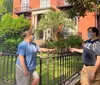 Two people one wearing a face mask are touching hands through a wrought-iron fence in front of a historic-looking red brick building with greenery