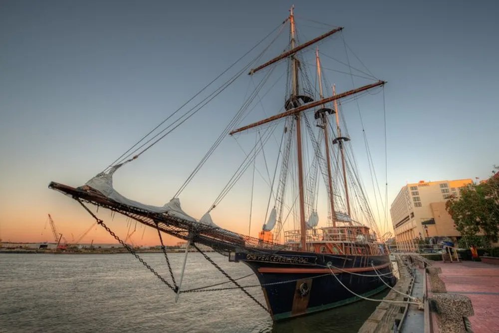 The image shows a tall ship docked at a peaceful harbor during sunset