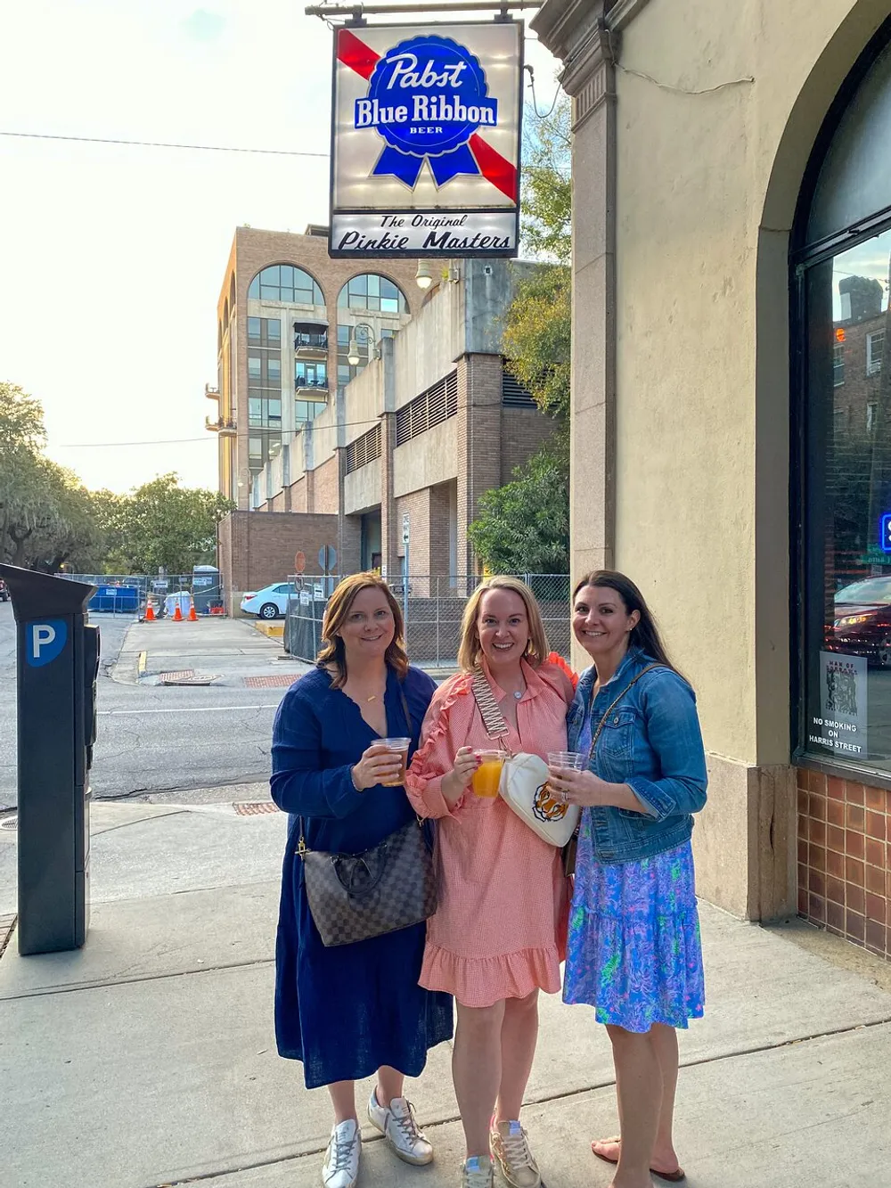 Three women are smiling for the camera while holding drinks on a sunny street corner under a Pabst Blue Ribbon beer sign