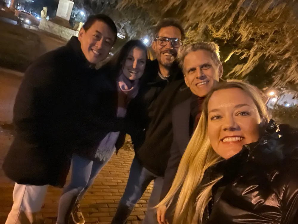 A group of five people are posing for a smiling selfie at night presumably enjoying a social gathering