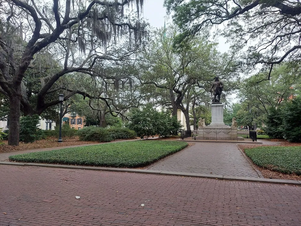 The image shows a peaceful urban park with a brick pathway leading to a statue surrounded by lush greenery and draped with Spanish moss-laden oak trees