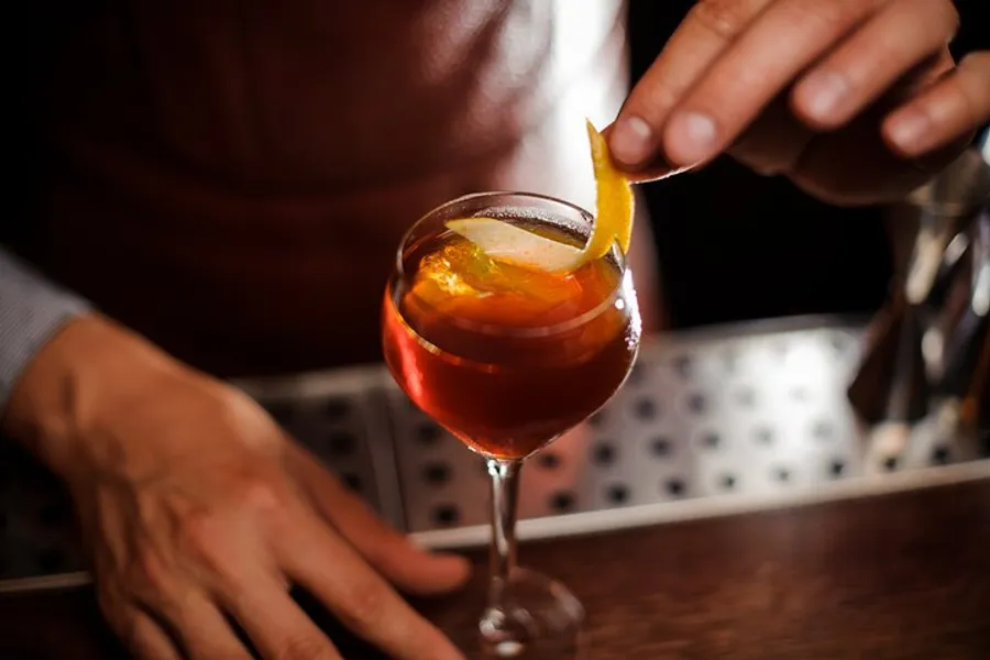 A bartender is garnishing a cocktail with an orange peel, presenting the drink with a flair on a bar counter.