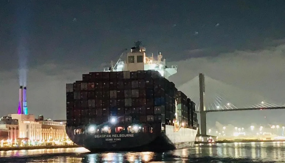 The image shows a large container ship named Seaspan Melbourne at night with a brightly lit multi-colored smokestack and a suspension bridge in the background