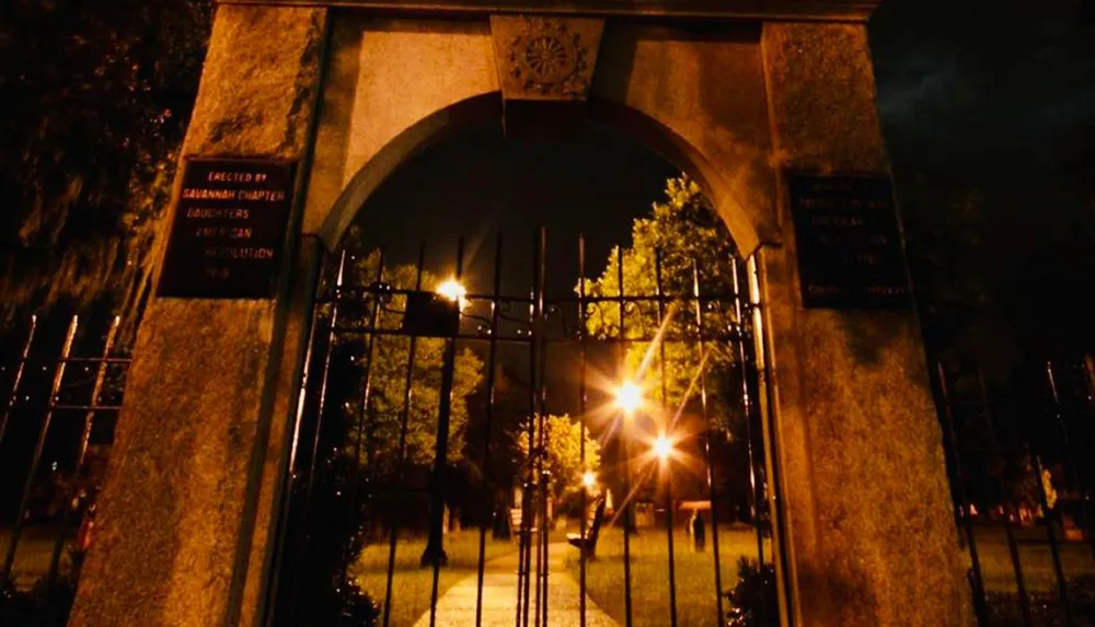 The image depicts a dimly lit scene of a gated entrance at night with commemorative plaques mounted on the adjacent pillars