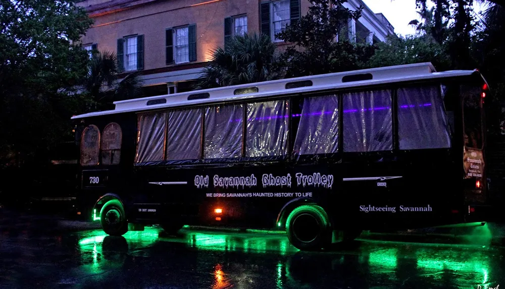 A black trolley with green and purple lights is parked on a wet street at dusk advertising the Old Savannah Ghost Trolley tour