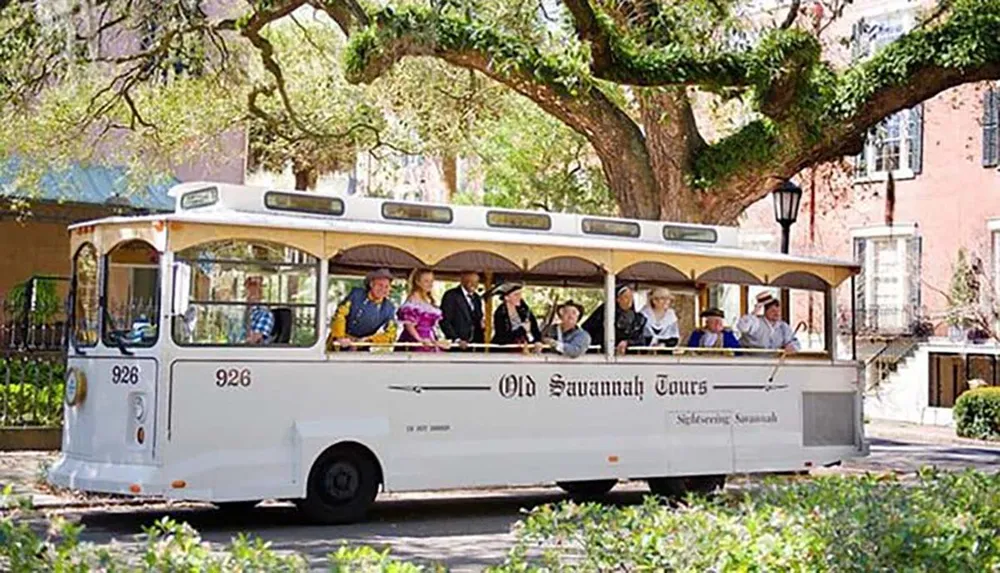 A group of people some dressed in period costumes are riding on a trolley labeled Old Savannah Tours through a picturesque street with lush trees