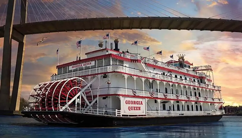 A large paddlewheel riverboat named the Georgia Queen is pictured on the water at dusk with a bridge overhead and a vibrant sky in the background