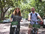A smiling man and woman are on a bicycle tour on a tree-lined street, with signs on their bikes that read 