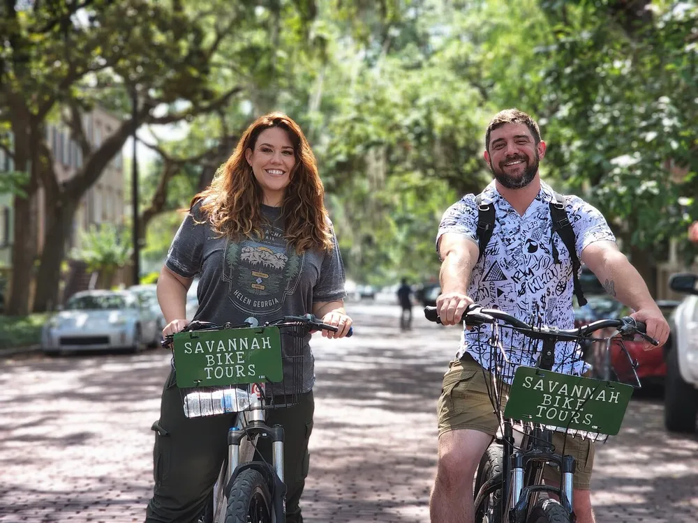 A smiling man and woman are on a bicycle tour on a tree-lined street with signs on their bikes that read Savannah Bike Tours