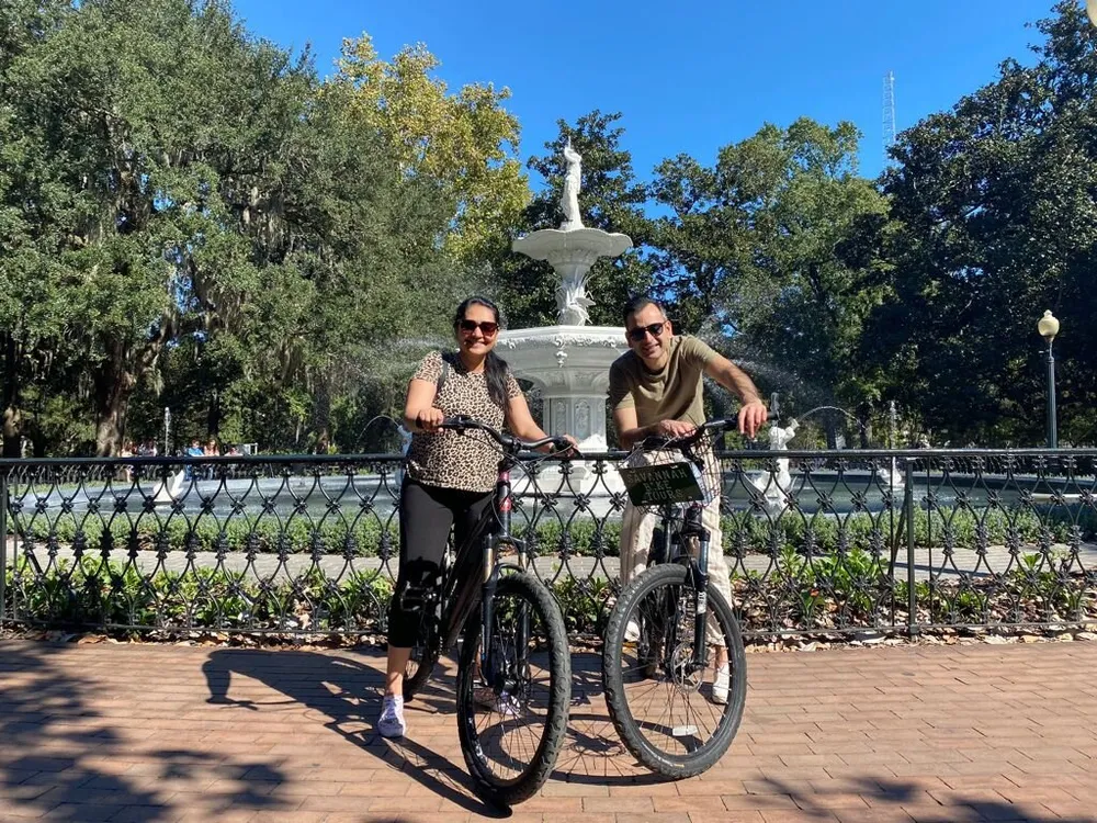 Two people are smiling and posing with bicycles in a sunny park with a fountain in the background