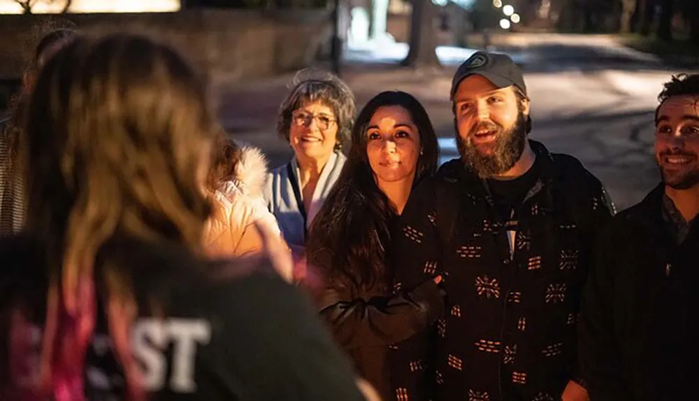 A group of smiling people are standing outside at night engaging in a lively conversation with someone whose back is facing the camera