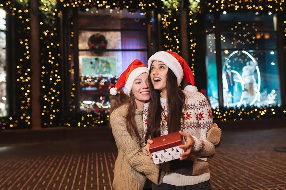 Two people wearing Santa hats are joyfully posing with a gift box against a festive illuminated background suggestive of holiday cheer
