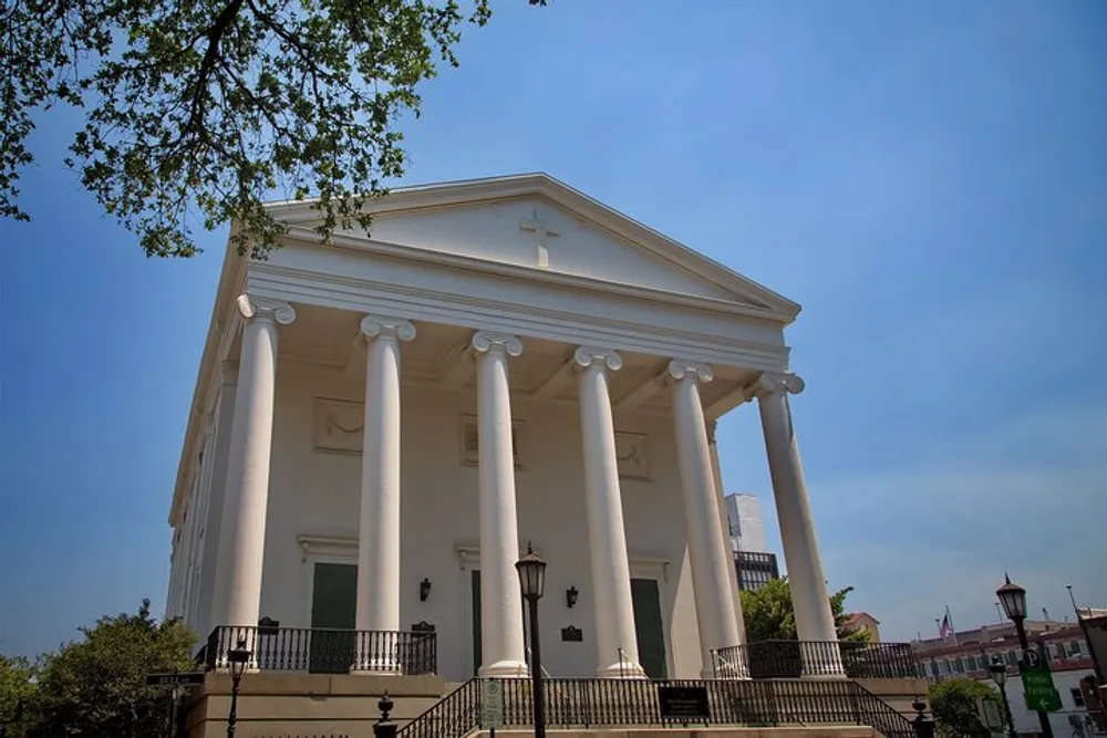 The image shows a neoclassical building with large white columns and a cross at the top suggesting it may be a church or another type of important historical edifice set against a blue sky with some foliage visible at the edge