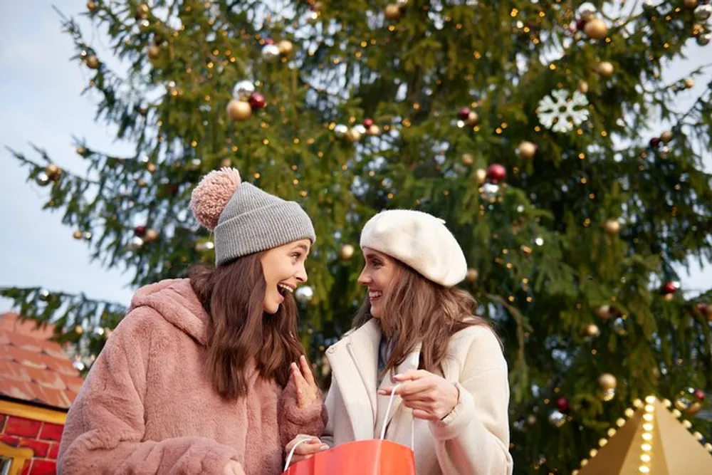 Two women are joyfully conversing against the backdrop of a festively decorated Christmas tree both wearing winter hats and holding a shopping bag